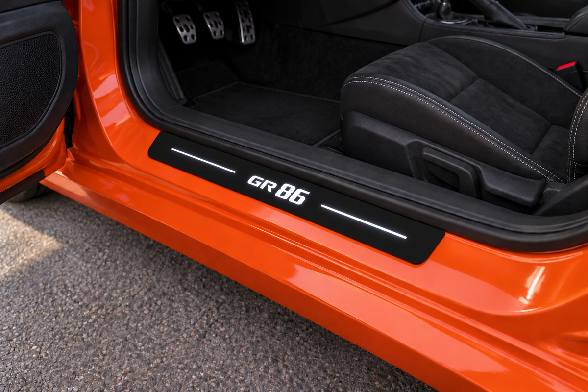 Toyota GR86 ZN8 Matte PRO sill visualization: a separate black plate on an exposed orange metal threshold directly against the rubber seal, with white GR86 lettering and two top light bars; edges remain unlit.