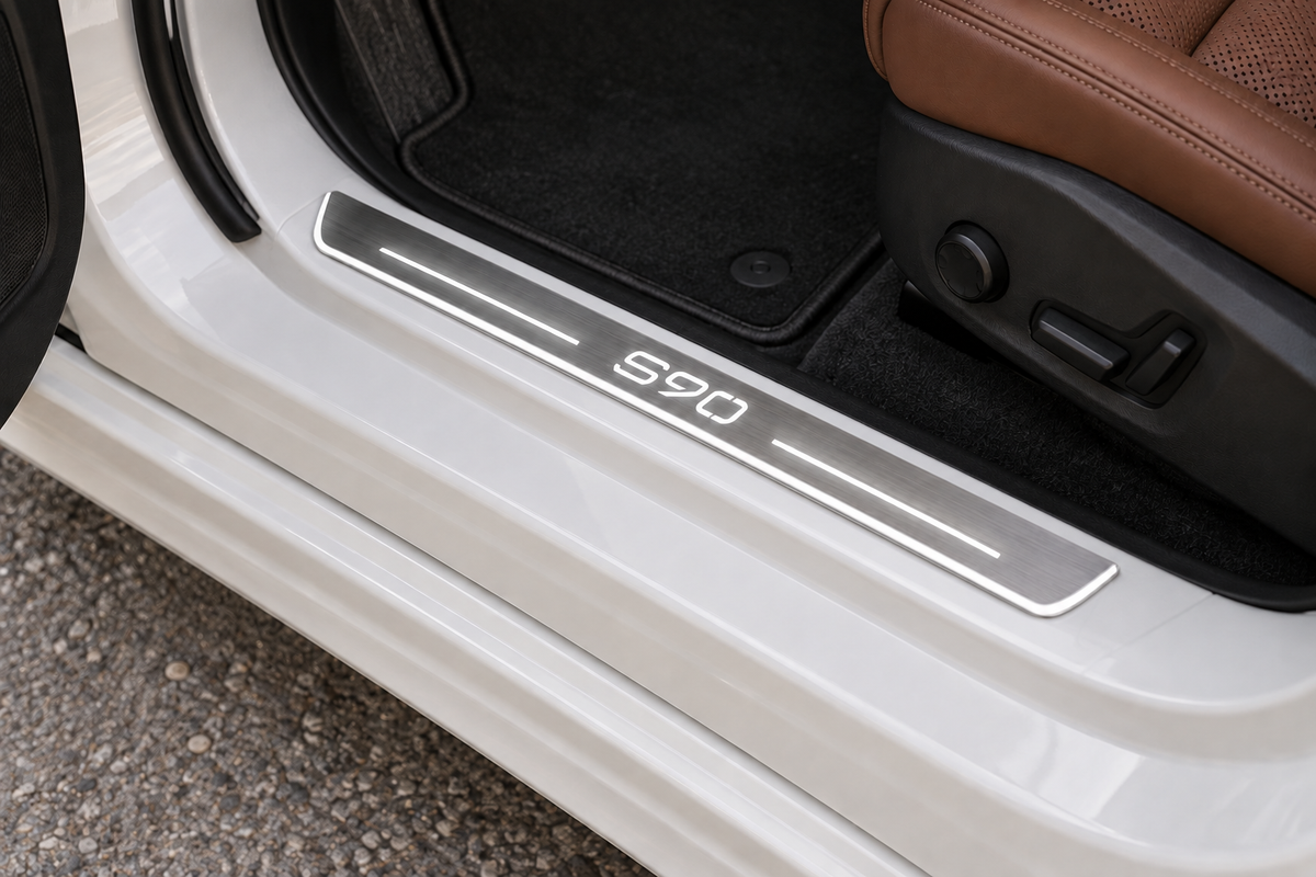 Volvo S90 stainless door sill visualization, seated directly on the white painted metal threshold with the factory plastic scuff cover removed; S90 lettering, top bars and stainless edge illumination.