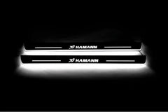Two long door sill plates with Hamann with winged emblems lettering and edges illuminated in white, straight-on view on a dark background.