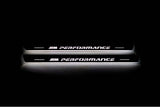 Two long door sill plates with M Performance lettering and edges illuminated in white, straight-on view on a dark background.