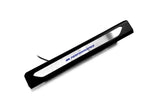 Black molded threshold trim with a silver insert bearing blue M Performance lettering and an attached wire, angled view.