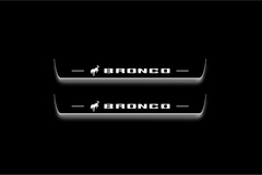Two long door sill plates with Bronco and a bucking horse emblem lettering and edges illuminated in white, straight-on view on a dark background.