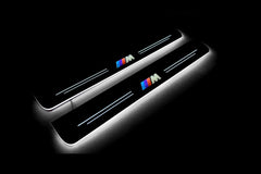 Two long door sill plates with a tricolor M emblem lettering and edges illuminated in white, angled view on a dark background.