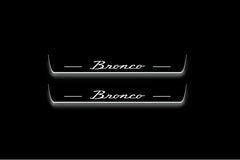 Two long door sill plates with Bronco in script lettering and edges illuminated in white, straight-on view on a dark background.