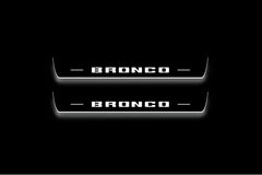 Two long door sill plates with Bronco in block lettering lettering and edges illuminated in white, straight-on view on a dark background.
