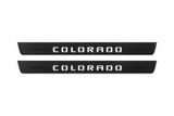 Catalogue illustration of two black Matte PRO front door sill plates with illuminated geometric COLORADO lettering on the upper surface. Decorative lines and edges are unlit; rear plates for FRONT+REAR are not pictured. Dimensions are selected for the Chevrolet Colorado II during manufacture.