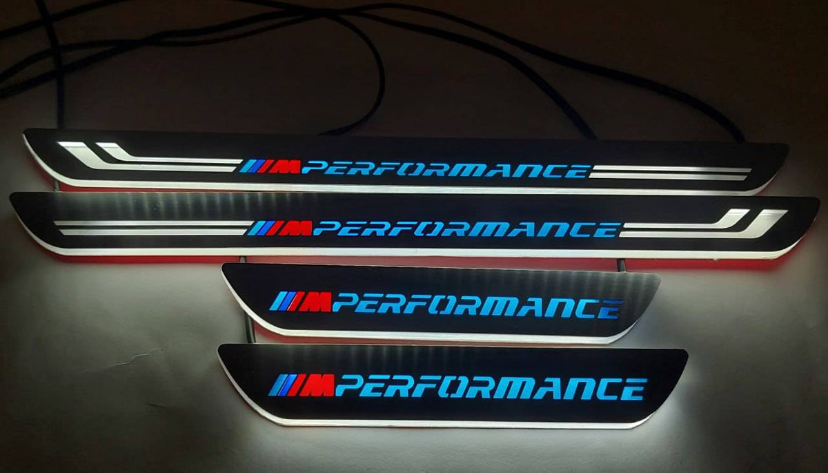 Four wired door sill plates with blue M Performance lettering, tricolor M emblems and white illuminated edges, straight-on view.