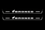 Product visualization: two flat Bronco front plates, one horse and bars on each. Black night background, unlit stainless face nearly hidden, white illuminated artwork, bars and real side/end edges with soft glow.