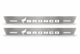 Product visualization: two Ford Bronco FRONT, exact one horse and stencil logo flat brushed stainless steel door sill plates with white LED artwork and edges, white background.