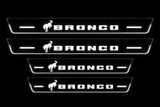 Front+Rear: four flat Stainless steel Bronco door sill plates, two long front and two shorter rear, one horse and BRONCO lettering per plate; white illuminated artwork, stripes and edges on a black background.