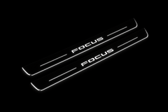 Product visualization: two exact focus-iv-diagonal flat Stainless front sill plates with original artwork, neutral white LEDs and own thin-edge geometry flat brushed stainless steel door sill plates with white LED artwork and edges, dark background.