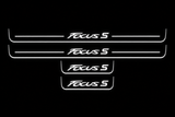 Product visualization: four original flat Focus S sill plates, two front with bars and two shorter rear without bars flat brushed stainless steel door sill plates with white LED artwork and edges, dark background.