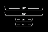 Product visualization: four flat Stainless Steel door-sill plates, two long front and two shorter rear plates. Original artwork and actual edge/end LED zones illuminated white on black; not an installation photograph.