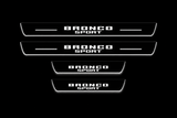 Product visualization: four flat Stainless Steel door-sill plates, two long front and two shorter rear plates. Original artwork and actual edge/end LED zones illuminated white on black; not an installation photograph.