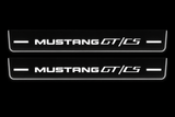 Product visualization: two flat Mustang GT/CS front sill plates, exact current v3 MUSTANG and GT/CS artwork. Black night background, unlit stainless face nearly hidden, white illuminated artwork, bars and real side/end edges with soft glow.