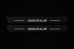 Catalogue illustration of two black Matte PRO front door sill plates with stylized white mach E 4 X lettering and thin horizontal lines illuminated only on top. Rear plates for FRONT+REAR are not pictured; dimensions are selected for the vehicle during manufacture.