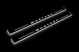 Product visualization: MUSTANG spaced letters, two flat source front plates flat brushed stainless steel door sill plates with white LED artwork and edges, dark background.
