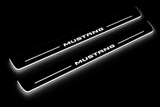 Product visualization: MUSTANG compact block lettering, two flat source front plates flat brushed stainless steel door sill plates with white LED artwork and edges, dark background.