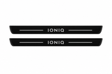 Catalogue illustration of two black Matte PRO front door sill plates with illuminated white IONIQ lettering and thin top light lines. The edges remain unlit. Rear plates for FRONT+REAR are not pictured; dimensions are selected for the vehicle during manufacture.