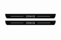 Catalogue illustration of two black Matte PRO front door sill plates with illuminated white IONIQ lettering and thin top light lines. The edges remain unlit. Rear plates for FRONT+REAR are not pictured; dimensions are selected for the vehicle during manufacture.
