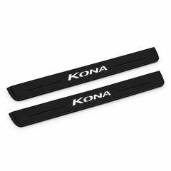 Catalogue illustration of two black Matte PRO front door sill plates with illuminated KONA lettering on the top surface. Edges are unlit; rear plates for FRONT+REAR are not pictured. Final dimensions are selected for Hyundai Kona OS during manufacture.