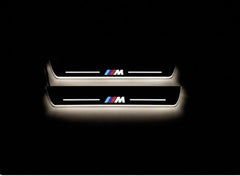 Two short dark door sill plates with tricolor M emblems and edges illuminated warm white on a black background.
