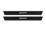 Catalogue illustration of two black Matte PRO front door sill plates with illuminated lowercase picanto lettering on top. Edges remain unlit. Rear plates for a FRONT+REAR set are not shown; final dimensions are chosen for the Kia Picanto JA during manufacture.