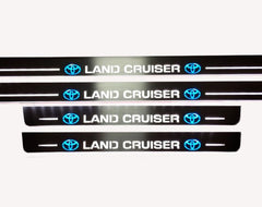 Close-up of white Land Cruiser lettering flanked by blue Toyota emblems on four reflective door sill plates.