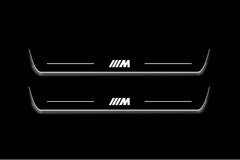 Two long door sill plates with a striped M emblem lettering and edges illuminated in white, straight-on view on a dark background.
