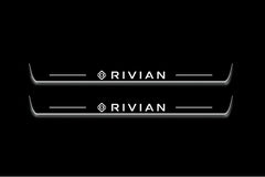 Two long door sill plates with Rivian with diamond emblems lettering and edges illuminated in white, straight-on view on a dark background.
