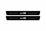 Catalogue illustration of two black Matte PRO front door sill plates with white GR86 lettering and thin horizontal lines on the top surface. Rear plates for FRONT+REAR are not pictured; final dimensions are selected for the vehicle during manufacture.
