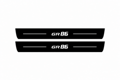 Catalogue illustration of two black Matte PRO front door sill plates with white GR86 lettering and thin horizontal lines on the top surface. Rear plates for FRONT+REAR are not pictured; final dimensions are selected for the vehicle during manufacture.