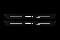 Catalogue illustration of two black Matte PRO front door sill plates with white TRUENO and lowercase italic edition lettering and thin lines illuminated only on the top surface. Rear plates for FRONT+REAR are not pictured; final dimensions are selected for the vehicle during manufacture.