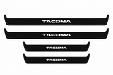Catalogue illustration of two long black Matte PRO front sill plates and two shorter rear plates with TACOMA lettering illuminated on top. Side edges unlit; vehicle-specific dimensions are not depicted. FRONT option includes front plates only.