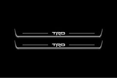 Two long door sill plates with TRD lettering and edges illuminated in white, straight-on view on a dark background.